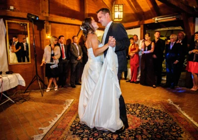 Ein Brautpaar tanzt auf einem dekorativen Teppich, umgeben von lachenden Gästen in einem holzgetäfelten Raum.