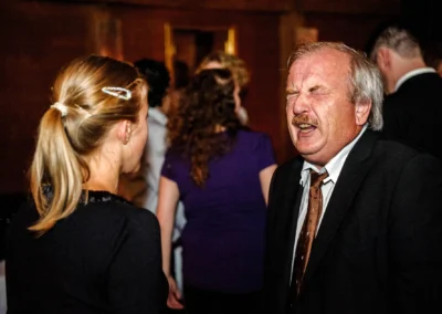 Eine Frau mit Haarspange im Gespräch mit einem Mann im Anzug, der die Augen geschlossen hat; im Hintergrund unscharfe Personen.