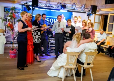 Gäste bei einer Hochzeit in Kreuth am Tegernsee singen für das Brautpaar, das entspannt auf Stühlen sitzt. Hochzeitsfoto.