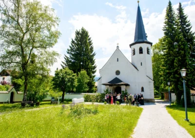Kirchliche Trauung in Garmisch mit Hochzeitsgaesten vor kleiner Kirche, umgeben von Baeumen und Gruenflaeche.