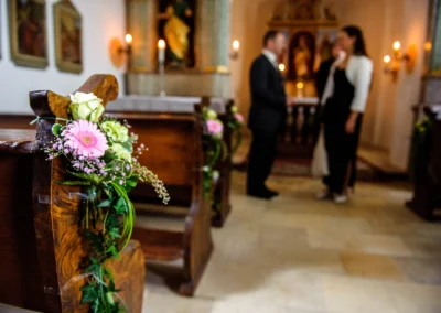 Blumengesteck auf Kirchenbank bei einer Hochzeit in Kranzberg, Paar im Hintergrund. Hochzeitsfotografie Oberbayern.