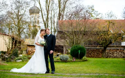 Brautpaar in der Natur; sie trägt ein weißes Kleid, er einen dunklen Anzug. Im Hintergrund Bäume und ein Gebäude mit Holzstapel.