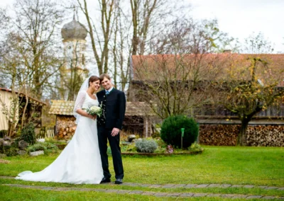 Brautpaar steht lächelnd in einem Garten, Kirche im Hintergrund, Hochzeitsfotografie in Holzkirchen südlich von Muenchen.