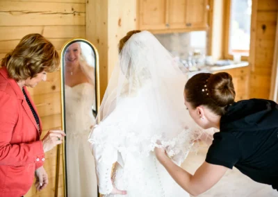 Brautkleid-Anprobe bei Hochzeit in Tegernsee. Zwei Frauen helfen der Braut mit Schleier vor dem Spiegel. Hochzeitsfoto.