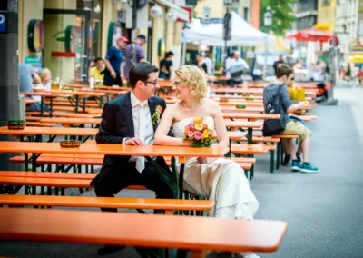 Brautpaar sitzt bei der Hochzeitsfotografie in Schwabing an einem Tisch im Freien und strahlt sich an.
