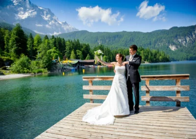 Brautpaar auf Steg am Eibsee mit Bergen im Hintergrund, Hochzeitsfoto bei sonnigem Wetter nahe Garmisch.