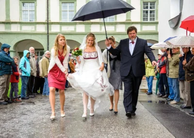 Braut schreitet mit Begleitung und Regenschirm in Kloster Benediktbeuern zur kirchlichen Trauung einer Hochzeit.