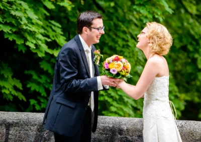Brautpaar lacht sich an, waehrend sie einen Blumenstrauss halten. Hochzeitsfoto in Schwabing von einem Fotografen.