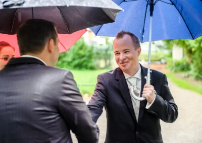 Bräutigam beim Handschlag mit Gast unter Regenschirmen bei kirchlicher Trauung in Pasing, Hochzeitsreportage.