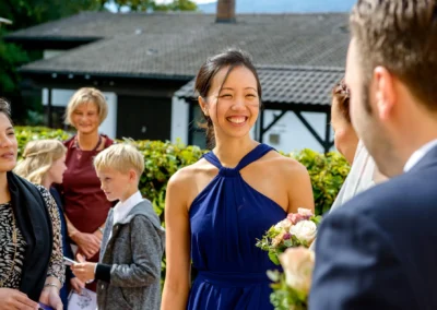 Lachende Frau in blauem Kleid haelt Blumenstrauss bei Hochzeitsreportage in Fischbachau. Hochzeitsfoto im Freien.