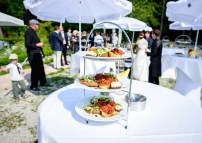Hochzeitsfoto am Starnberger See: Etagere mit Speisen im Vordergrund, Gaeste und Brautpaar im Hintergrund.