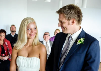 Brautpaar laechelt sich bei standesamtlicher Trauung in Bernried an, begleitet von Gaesten im Hintergrund. Hochzeitserinnerung.