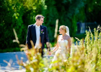 Brautpaar in eleganter Hochzeitskleidung spaziert Hand in Hand in Taufkirchen, umgeben von grünem Laub.
