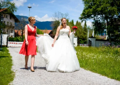 Braut im weissen Kleid mit Blumenstrauss und Begleiterin in rotem Kleid auf einem Pfad bei einer Hochzeit in Garmisch.