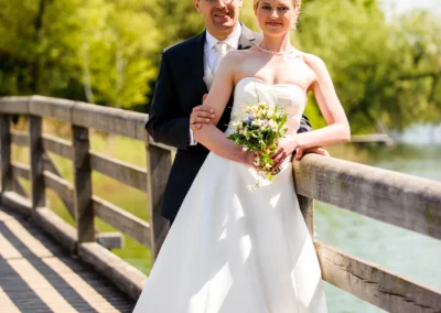 Brautpaar steht auf Holzbrücke am Wasser, Hochzeitsfotografie Kloster Seeon im Chiemgau, strahlender Sonnenschein.