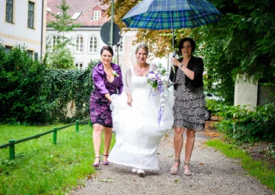 Braut mit Blumenstrauss unter Regenschirm, begleitet von zwei Frauen, auf dem Weg zur kirchlichen Trauung in Pasing.