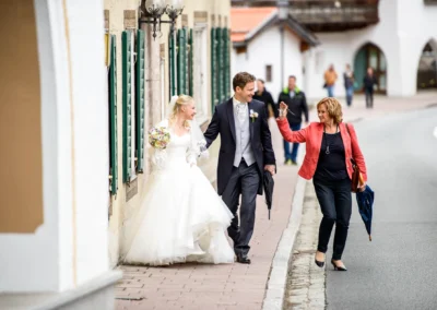 Brautpaar in Tegernsee spaziert lachend auf der Strasse, begleitet von einer Frau mit Schirm. Hochzeitsfotografie.