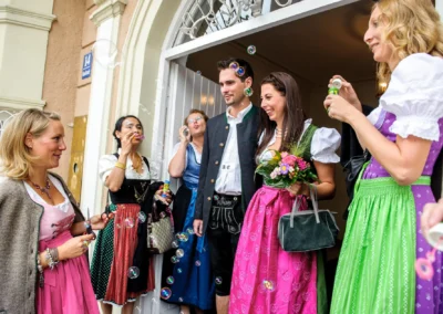 Gruppenszene mit lachenden Hochzeitsgaesten in traditioneller Tracht beim Seifenblasen nach Trauung im Standesamt Schwabing.
