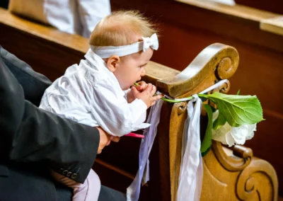 Kleinkind bei kirchlicher Hochzeit in Waakirchen, neugierig an Holzbank mit Blumenarrangement. Hochzeitsreportage.