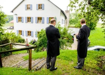 Zwei Maenner in Anzuegen in einem Garten, Gebaeude im Hintergrund. Hochzeitsfotografie bei Freising, kirchliche Trauung.