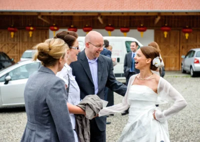 Braut in weissem Kleid lacht mit Gaesten vor Holzgebäude bei einer Hochzeit auf Gut Thurnsberg nordlich von Muenchen.