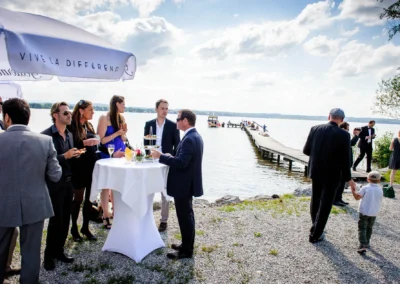Gäste bei Hochzeit am Starnberger See im Freien. Menschen unterhalten sich an Stehtischen. Hochzeitsreportage.