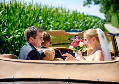 Brautpaar im Auto lacht, Braut hält Blumenstrauß, Hochzeitsfoto in Taufkirchen bei München.
