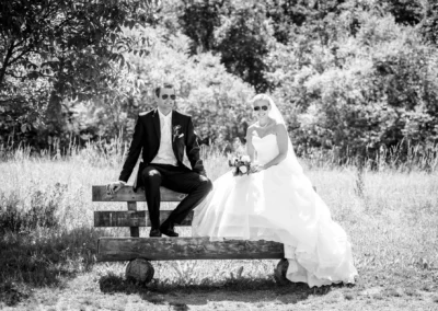 Brautpaar mit Sonnenbrillen auf Holzbank in ländlicher Umgebung, Taufkirchen bei München, Hochzeitsfoto.