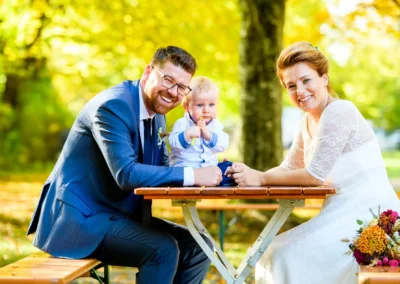 Brautpaar mit Kind an Holztisch im Gruenwalder Forst, Hochzeitsfotografie lebendig und natuerlich.