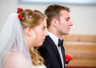 Brautpaar waehrend der kirchlichen Trauung in Garmisch, Braut mit Schleier, Bräutigam mit roser Rose. Hochzeitsfotografie.