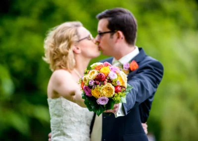 Brautpaar mit Blumenstrauss in Schwabing bei Hochzeit, festgehalten von Hochzeitsfotograf in gruenem Gartenumfeld.
