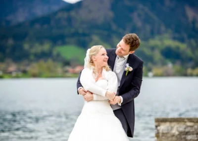 Brautpaarfoto am Tegernsee, laechelnd am Wasser. Hochzeit mit Bergen im Hintergrund. Hochzeitsfotografie in Bayern.