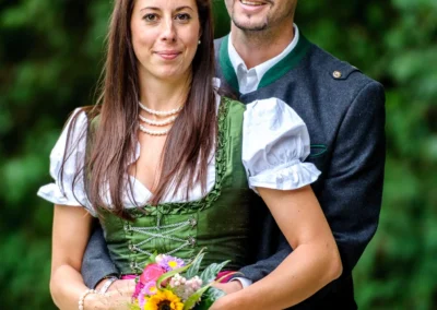 Brautpaar bei Hochzeitsportraits im Englischen Garten in Muenchen mit Blumenstrauss und traditionellen Trachten.