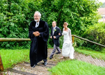 Brautpaar mit Geistlichem bei kirchlicher Trauung auf Stufen, umgeben von Gruen. Hochzeitsfotografie bei Freising.