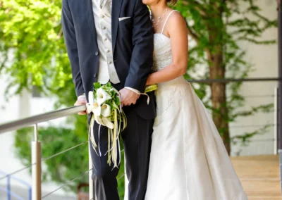 Brautpaar bei Hochzeit am Eibsee, Hochzeitsfotografie auf Terrasse mit gruenem Hintergrund, elegante Kleidung und Blumenstrauss.