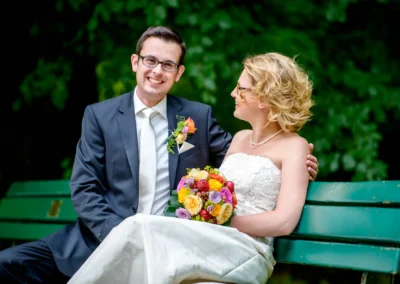 Brautpaar auf Bank im Englischen Garten München, lachend, mit Blumenstrauss. Hochzeitsfoto, Hochzeitsfotograf in Schwabing.
