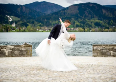 Brautpaar tanzt vor malerischer Kulisse am Tegernsee. Hochzeitsfoto mit Bergblick und verliebtem Paar.