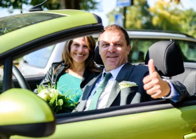 Glückliches Paar im Auto bei einer Hochzeitsreportage in München, lächelnd mit Blumenstrauß. Hochzeit in Obermenzing.