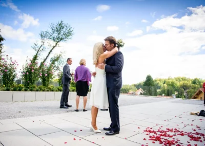Brautpaar bei der standesamtlichen Trauung am Starnberger See, im Hintergrund Gaeste und Bluetenblaetter.