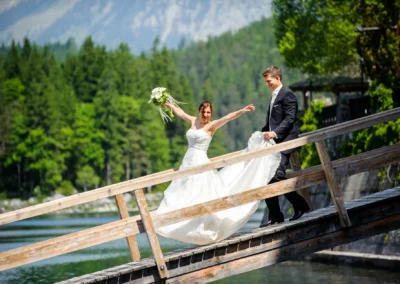 Brautpaar auf einer Holzbrücke am Eibsee, lachend in Hochzeitskleidung. Hochzeitsfotograf für natuerliche Momente.