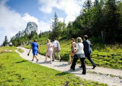 Hochzeitsgaeste spazieren auf einem Pfad bei der Hochzeit nahe Kampenwand. Berge und Baeume im Hintergrund.