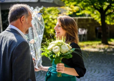 Frau mit Blumenstrauss und Mann bei kirchlicher Trauung in Muenchen. Hochzeitsreportage bei Sonnenschein im Freien.