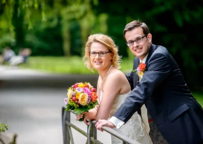 Brautpaarfoto im Englischen Garten München, laechelnd an einem Geländer. Hochzeitsfotografie in natuerlicher Umgebung.