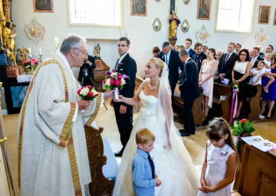Braut uebergibt Blumen an Priester waehrend kirchlicher Trauung in Taufkirchen nahe Muenchen, umgeben von Gaesten.