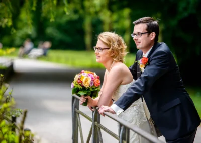 Brautpaar lehnt an einem Geländer im Englischen Garten München. Hochzeitsfoto mit Blumenstrauss.