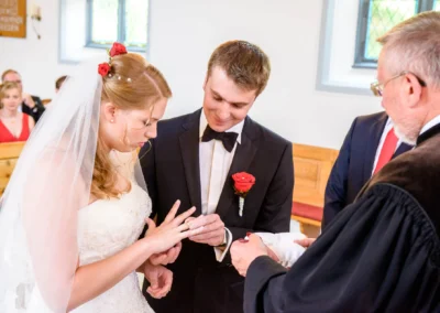 Brautpaar bei kirchlicher Trauung in Garmisch. Braut steckt Ring an, traegt Schleier mit roten Blumen. Hochzeitsfotograf.