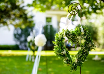 Herzfoermiger Kranz aus Gruen mit weissen Schleifen bei Hochzeit am Tegernsee. Hochzeitsfoto zeigt Gartenkulisse.