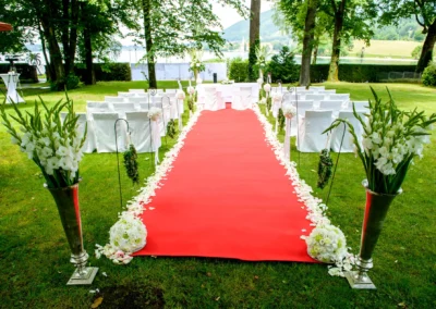 Roter Teppich und Blumenarrangement bei Hochzeit am Tegernsee. Weiße Stühle im Freien. Hochzeitsfoto.