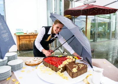 Hochzeitsfotografie: Kellnerin mit Regenschirm neben Hochzeitstorte bei Regen am Tegernsee. Geschirr im Hintergrund.