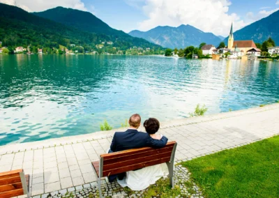 Brautpaar sitzt auf Bank mit Blick auf Tegernsee und Berge. Hochzeitsfoto bei Rottach Egern am Tegernsee.